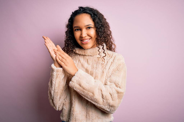 Jonge mooie vrouw met krullend haar dragen casual trui staan over roze achtergrond klappen en applaudisseren gelukkig en vrolijk, glimlachende trotse handen samen - Foto, afbeelding