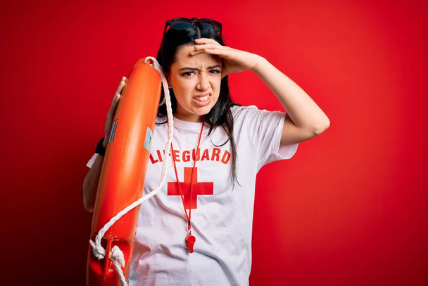 Young lifeguard woman wearing secury guard equipent holding rescue float over red background stressed with hand on head, shocked with shame and surprise face, angry and frustrated. Fear and upset for mistake. - Photo, Image