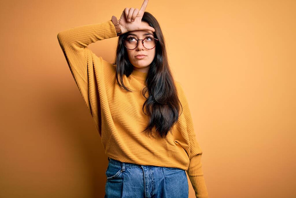 jonge brunette vrouw dragen bril en casual trui over geel geïsoleerde achtergrond maken plezier van mensen met vingers op het voorhoofd doen verliezer gebaar spotten en beledigend. - Foto, afbeelding