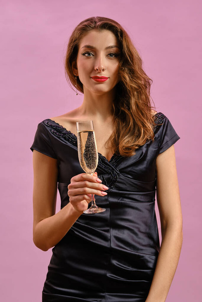 Brünette Frau im schwarzen Kleid. Sie hält ein Glas Champagner in der Hand und lächelt, posiert auf rosa Hintergrund. Mode und Schönheit. Nahaufnahme, Kopierraum - Foto, Bild