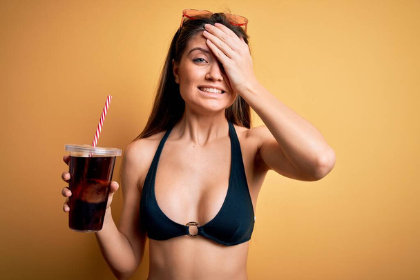 Young beautiful woman with blue eyes on vacation wearing bikini drinking cola beverage stressed with hand on head, shocked with shame and surprise face, angry and frustrated. Fear and upset for mistake. - Foto, Imagem