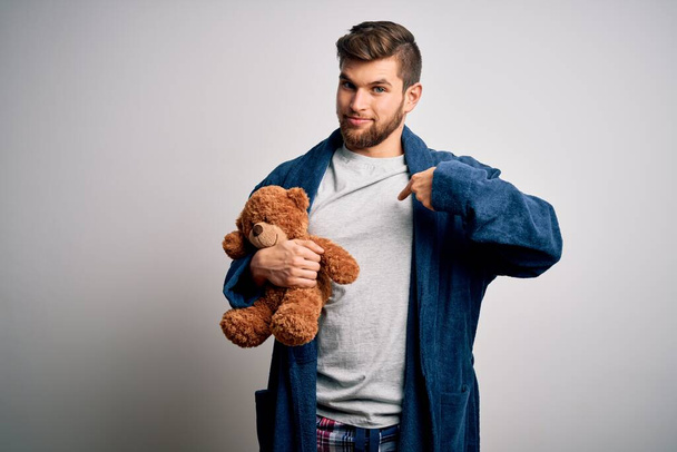 Young blond man with beard and blue eyes wearing pajama holding teddy bear with surprise face pointing finger to himself - Fotó, kép