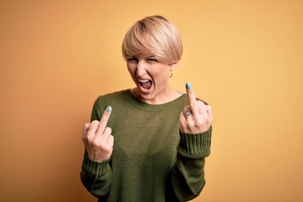 Young blonde woman with modern short hair wearing casual sweater over yellow background Showing middle finger doing fuck you bad expression, provocation and rude attitude. Screaming excited - Zdjęcie, obraz