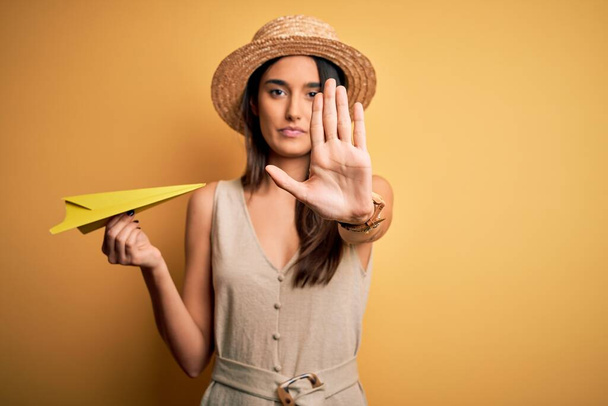 Молодая красивая брюнетка женщина в шляпе держа бумажный самолет над желтым фоном с открытой рукой делает знак стоп с серьезным и уверенным выражением, защитный жест
 - Фото, изображение