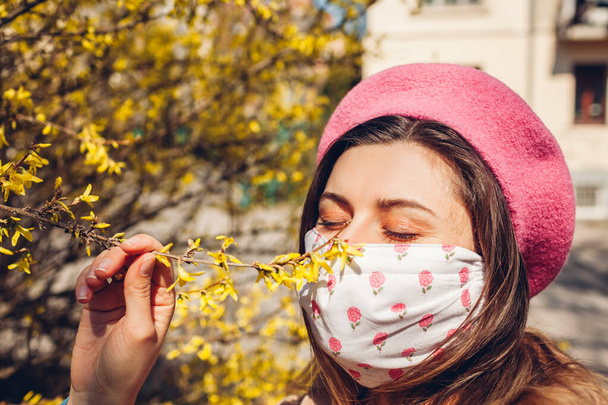 Coronavirus covid-19 salgını sırasında kadın açık havada tekrar kullanılabilir maske takıyor. Kız bahar çiçeklerinin kokusunu alıyor. Güvende kal, olumlu. Bahar modası - Fotoğraf, Görsel