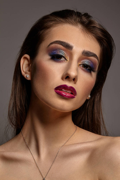 Brunette naked girl in jewelry posing on gray studio background. Luxury makeup. Colorful eyeshadow, long eyelashes, glossy red lips. Close up - Photo, Image