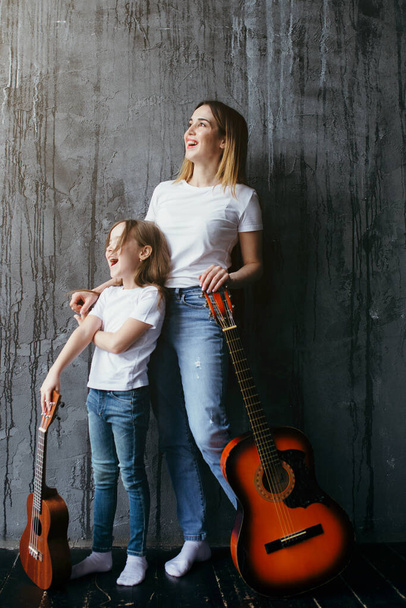 Mother teaches daughter to play the ukulele and guitar. Attractive mother and daughter look out the window at the sunset. - Фото, зображення