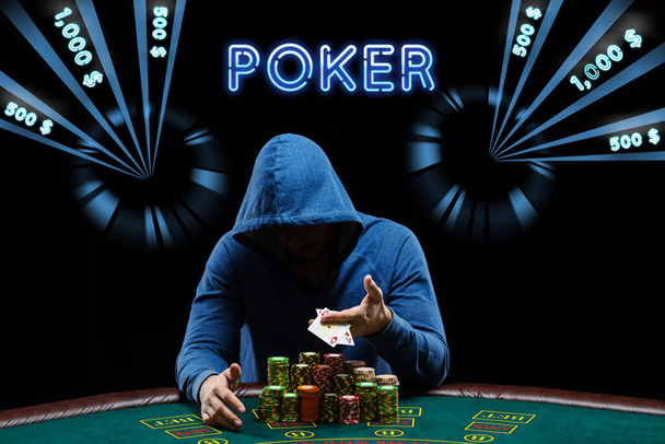 Mann in blauem Kapuzenpullover, der Asse zeigt, sitzt am Spieltisch mit Stapeln von Chips. Schwarzer Hintergrund mit neonfarbenen Dollarbeträgen, Beschriftungspoker. Casino - Foto, Bild