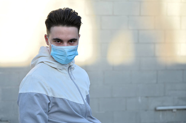 Portrait of a Caucasian young man in a medical mask outdoors in the city on a gray background on a sunny day - Photo, image