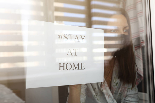 jonge vrouw in een medisch masker met papier met woorden thuis blijven achter een raam glas thuis. concept van quarantaine van de coronaviruspandemie, covid 19 - Foto, afbeelding