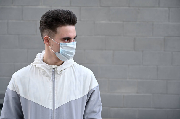 Photo of a Caucasian young brunette man in a medical protective mask in a light jacket outdoors against a gray wall in the city on a sunny day - Фото, изображение