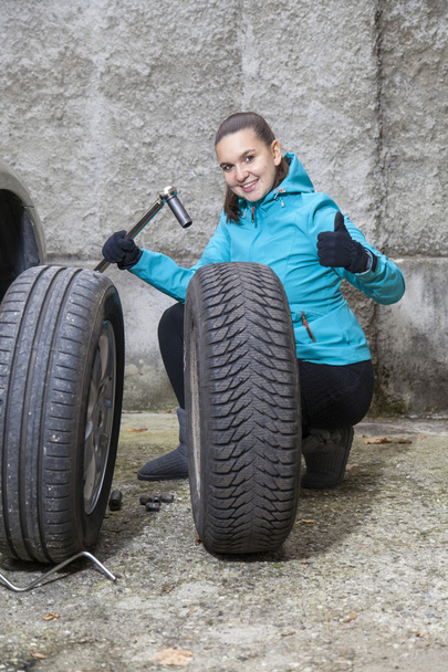 Nuori hymyilevä nainen kuljettaja korvaa renkaat, osoittaa OK merkki
 - Valokuva, kuva