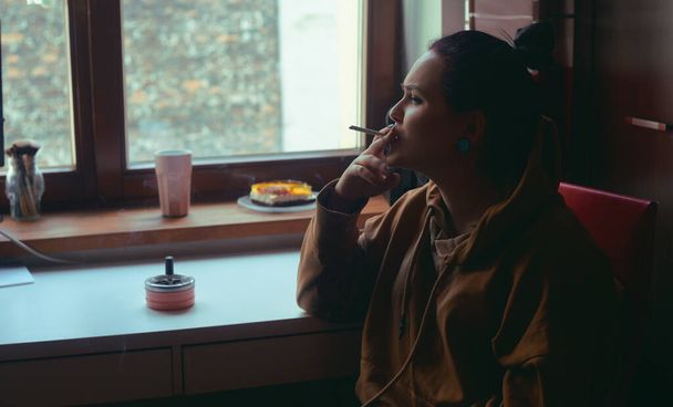 Evdeki kız oturur ve sigara içer, pencereden dışarı bakar. Yalnız kız pencerede sigara içiyor.. - Fotoğraf, Görsel