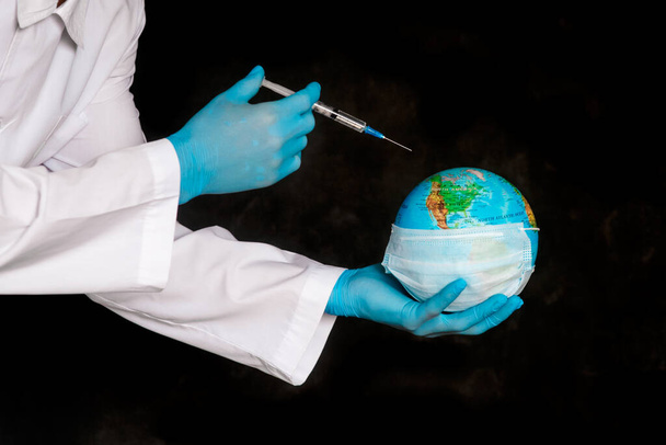 Globe earth in a medical mask and a syringe in the hands of the doctor close-up on a black background . The concept of the pandemic and the Coronavirus epidemic covid-19. - Photo, Image