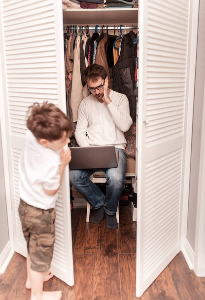 Busy father trying to find a quiet place in a wardrobe  to hide from children for a work at the laptop. Stay home concept. Quarantine. Freelancer. - Фото, изображение