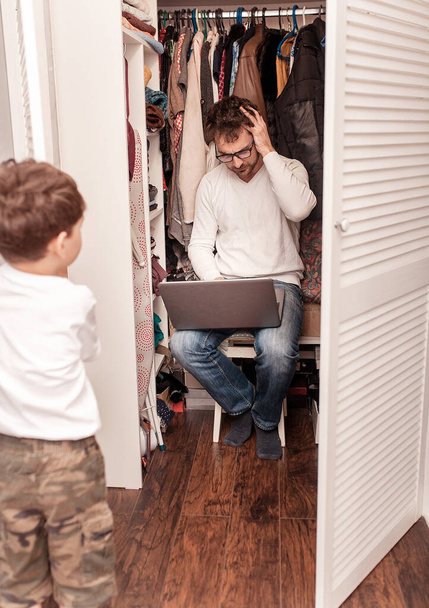 Busy father trying to find a quiet place in a wardrobe  to hide from children for a work at the laptop. Stay home concept. Quarantine. Freelancer. - Foto, Bild