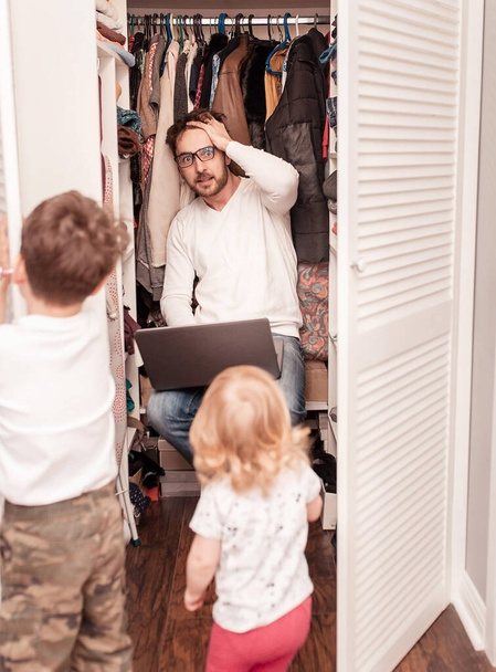 Busy father trying to find a quiet place in a wardrobe  to hide from children for a work at the laptop. Stay home concept. Quarantine. Freelancer. - Φωτογραφία, εικόνα
