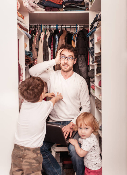 Busy father trying to find a quiet place in a wardrobe  to hide from children for a work at the laptop. Stay home concept. Quarantine. Freelancer. - Photo, image