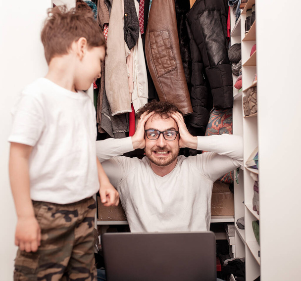 Busy father trying to find a quiet place in a wardrobe between clothes  to hide from children for a work at the laptop. Work from home. Home office. Quarantine. Freelancer. - Valokuva, kuva