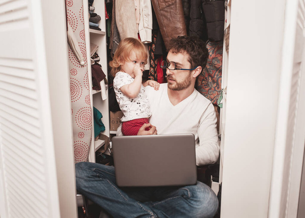 Busy father trying to find a quiet place in a wardrobe between clothes  to hide from children for a work at the laptop. Work from home. Home office. Quarantine. Freelancer. - Zdjęcie, obraz