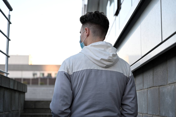Portrait of a Caucasian young brunette man in a light jacket outdoors on the background of the stairs of a building in the city. Model in a protective medical mask. Back view. - Fotografie, Obrázek