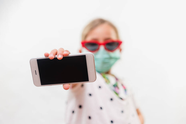 Little blond girl wearing disposable mask and sunglasses holding mobile phone over white background - Valokuva, kuva