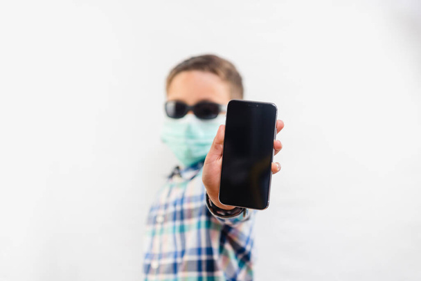 Young boy wearing disposable mask and sunglasses holding mobile phone over white background - 写真・画像