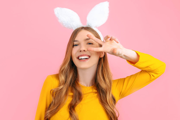 happy Easter. Smiling stylish woman in Easter Bunny ears, shows peace gesture, two fingers, on isolated pink background - Φωτογραφία, εικόνα