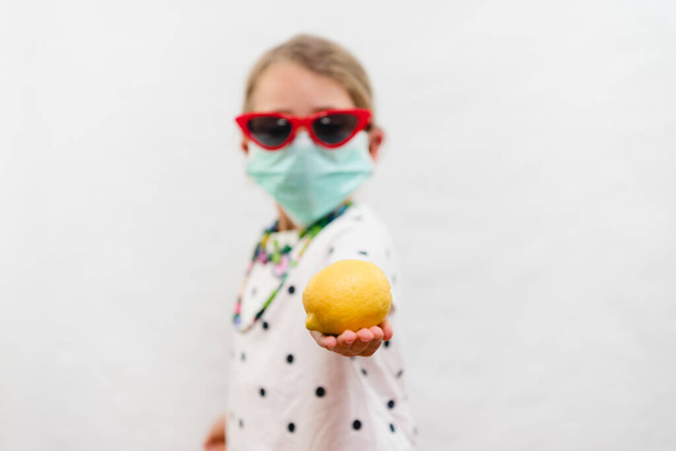 Kleines blondes Mädchen mit Einwegmaske und Sonnenbrille mit gelber Zitronen-Vitamin-C-Quelle vor weißem Hintergrund - Foto, Bild