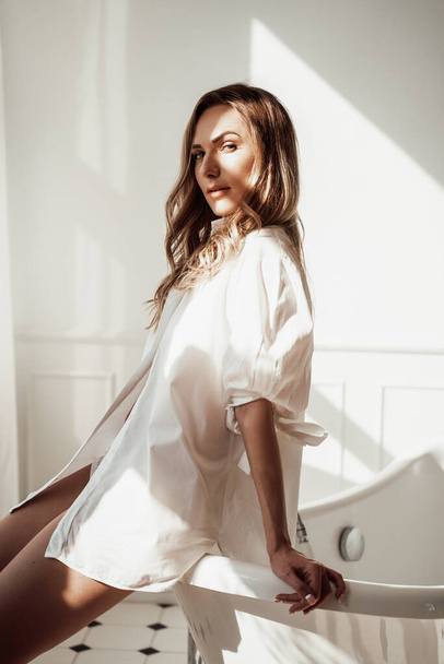 Sexy und sinnliche brünette Frau sitzt in einem hellen Badezimmer mit weißem Hemd und Dessous - Foto, Bild
