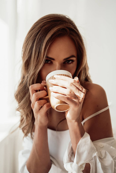 Graceful young woman in lingerie taking a sip from a cup of coffee over the white background - Valokuva, kuva