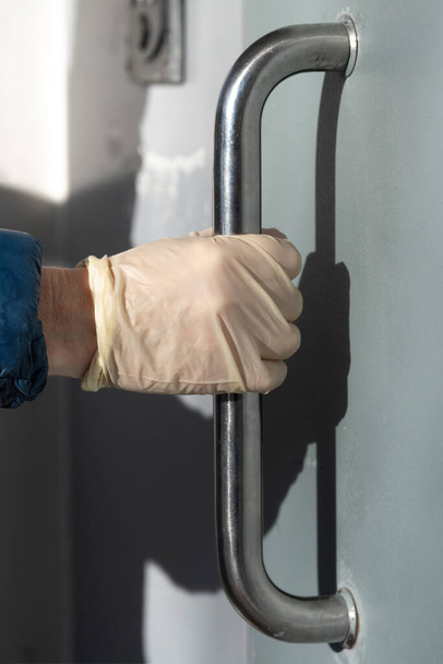 Hand in een beschermende handschoen om te voorkomen dat coronavirus infectie opent de deur door het metalen handvat - Foto, afbeelding