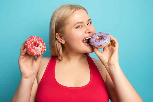 Fat girl eats sweet instead of do gym. Cyan background - Photo, Image