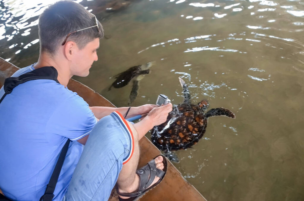 Młody brunetka facet z aparatem wypuszcza żółwia do wody, ochotnik ratuje żółwie, ochrony zwierząt, chłopiec robi zdjęcia żółwia. ratowanie zwierząt Sea Conservation Research Project Center w Bento - Zdjęcie, obraz