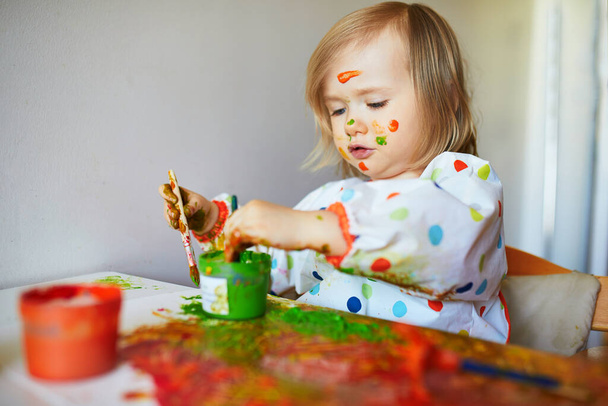 Adorable little girl with paint on her face painting with brushes and fingers at home, in kindergaten or preschool. Creative games for kids. Stay at home entertainment - Foto, Imagen