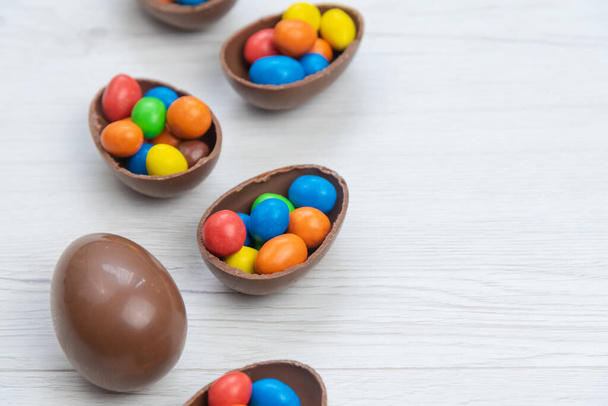 Chocolate Easter eggs and chocolate eggs filled with sweet and colorful candies, on white wood table. Copy space. - 写真・画像