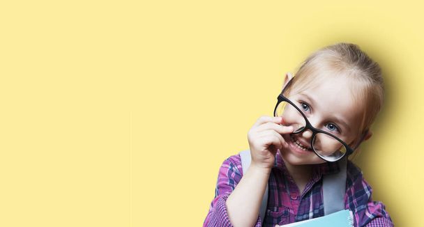 Aranyos szőke lány szemüvegben, hátizsákkal, narancssárga háttérrel. Óvodáskort tanít. Banner másolási hellyel - Fotó, kép