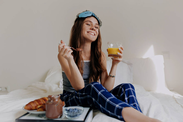 Ικανοποιημένο χαλαρό κορίτσι με μακριά κυματιστά μαλλιά που φοράει πιτζάμες κάθεται στο κρεβάτι με υπέροχο χαμόγελο και κλειστά μάτια και τρώει πρωινό στο κρεβάτι με κρουασάν και χυμό. - Φωτογραφία, εικόνα