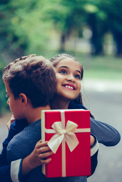 Šťastný chlapeček dává usměvavé dívce dárkovou krabici, která dělá překvapení. Šťastná holčička drží dárek v červené krabici a usmívá se při objímání svého chlapce - přítel. - Fotografie, Obrázek