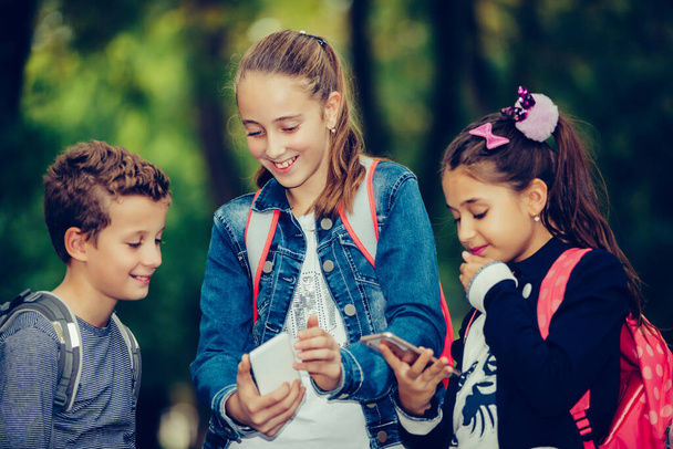 Ritratto all'aperto di ragazze positive e ragazzo che gioca con i telefoni dopo la scuola mentre cammina attraverso il parco
. - Foto, immagini