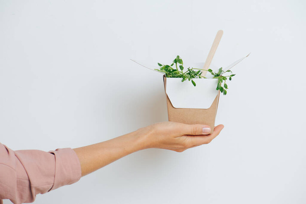 Woman hand holdig paper box full of pea strouts with bamboo fork sticking from it. Over white background. Disposable, compostable eco-friendly products. Side view. - 写真・画像