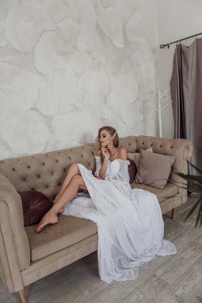 Fashion bride in a fashionable white dress with her hair and long earrings sitting on a modern sofa - Foto, Bild