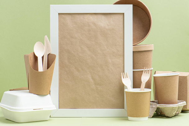 Frame in between of eco-friendly disposable bamboo cutlery, paper and sugarcane containers for food and drinks over green background. Side view on composition. Copy space. - 写真・画像