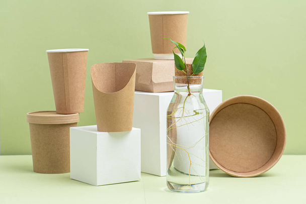 Eco-friendly disposable paper containers for food and drinks over green background. Side view on composition. Some objects placed on white cube pedestals, some placed on side. Plant branch in bottle. - Фото, изображение