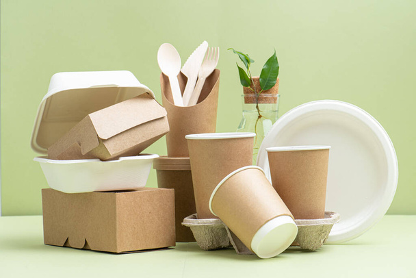 Eco-friendly disposable bamboo cutlery, paper and sugarcane containers for food and drinks over green background. Side view on composition. Plant branch in bottle behind. - Φωτογραφία, εικόνα