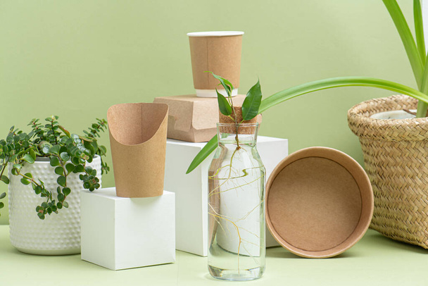Eco-friendly disposable paper containers for food and drinks over green background. Side view on composition. Some objects placed on white cube pedestals, some placed on side along with some plants. - Photo, image