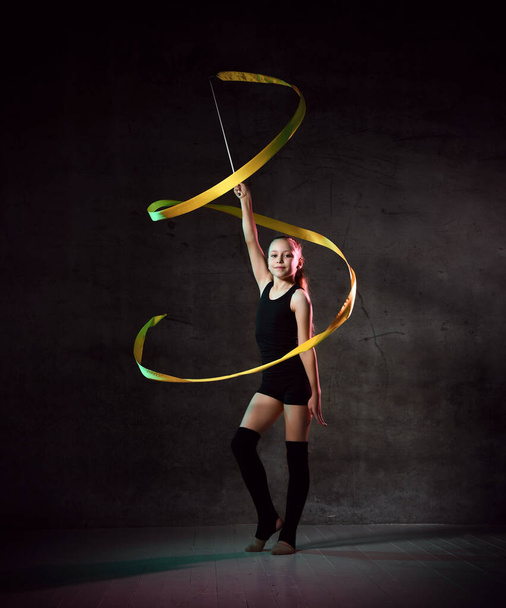 portrait of beautiful young brunette woman gymnast training calisthenics exercise with yellow ribbon on dark smoke studio background. - 写真・画像