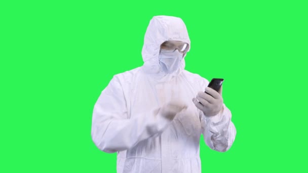 A man in a mask and a protective suit is talking via video calling on a smartphone while standing against a green background. - Video