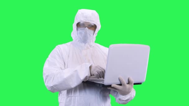 A man in a mask and glasses with a protective suit on a green background, looks around and enters data into a laptop - Video, Çekim