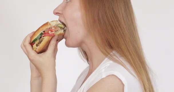Piękny głodny kobieta zjada duży burger i uśmiecha się odizolowany na białym tle - Materiał filmowy, wideo